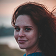 Close-up headshot of a young woman with reddish-brown, curly hair smiling slightly, with a soft outdoor background at sunset.